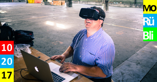Ein Mann sitzt in einer Halle an einem Laptop und trägt eine VR Brille.