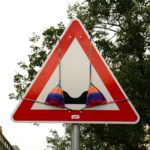 Straßenschild, das vor unebener Fahrband warnt, trägt vor den zwei stilisierten Hügeln einen bunten Bikini.