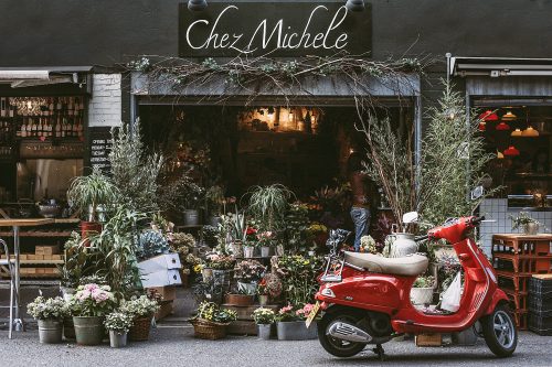 Pflanzenladen mit vielen, gestapelten Topfpflanzen. Eine rote Vespa rechts. Schild "Chez Michele".
