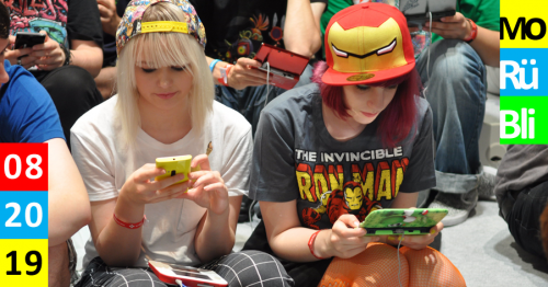 Titelbild Monatsrückblick. Eine Gruppe Menschen. Alle spielen am Nintendo DS. Zwei junge Frauen im Fokus. Beide tragen Fan-Merchandise. Beide spielen am Handy oder dem Nintendo DS.